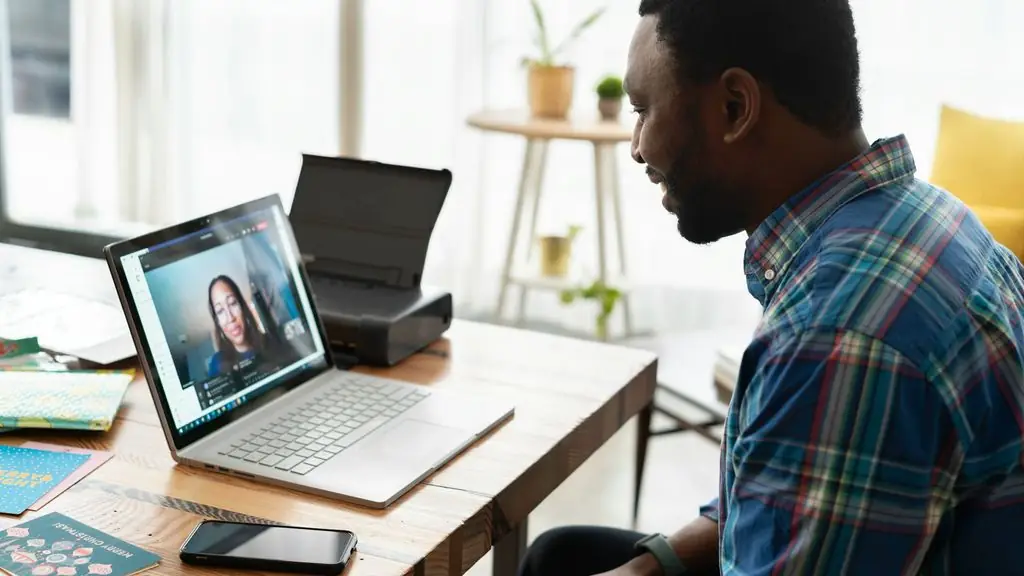 A man in a plaid shirt smiling at a laptop on his desk, where a woman appears on a video call, a phone and a few cards beside it.